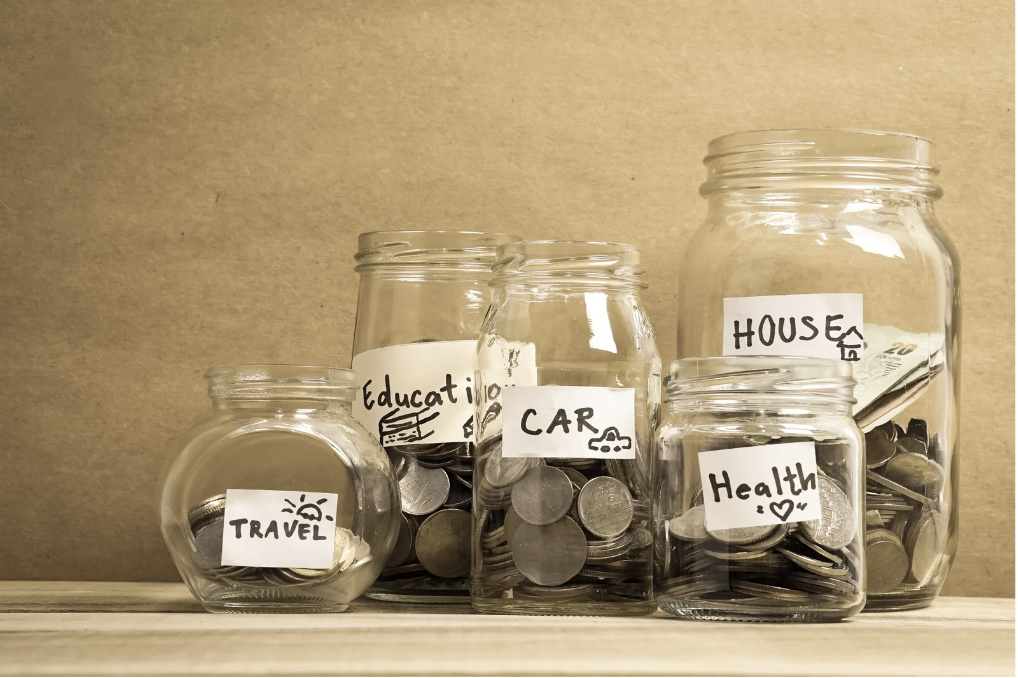 A picture of various size clear class jars filled with coins and labeled with white paper and black pen- travel, health, house, care, education